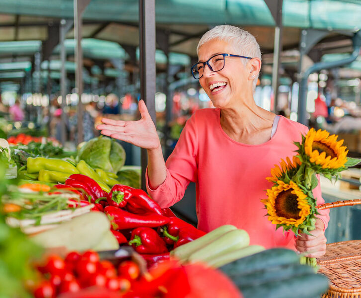 A senior woman at a farmer's market holding a basket and bouquet of sunflowers