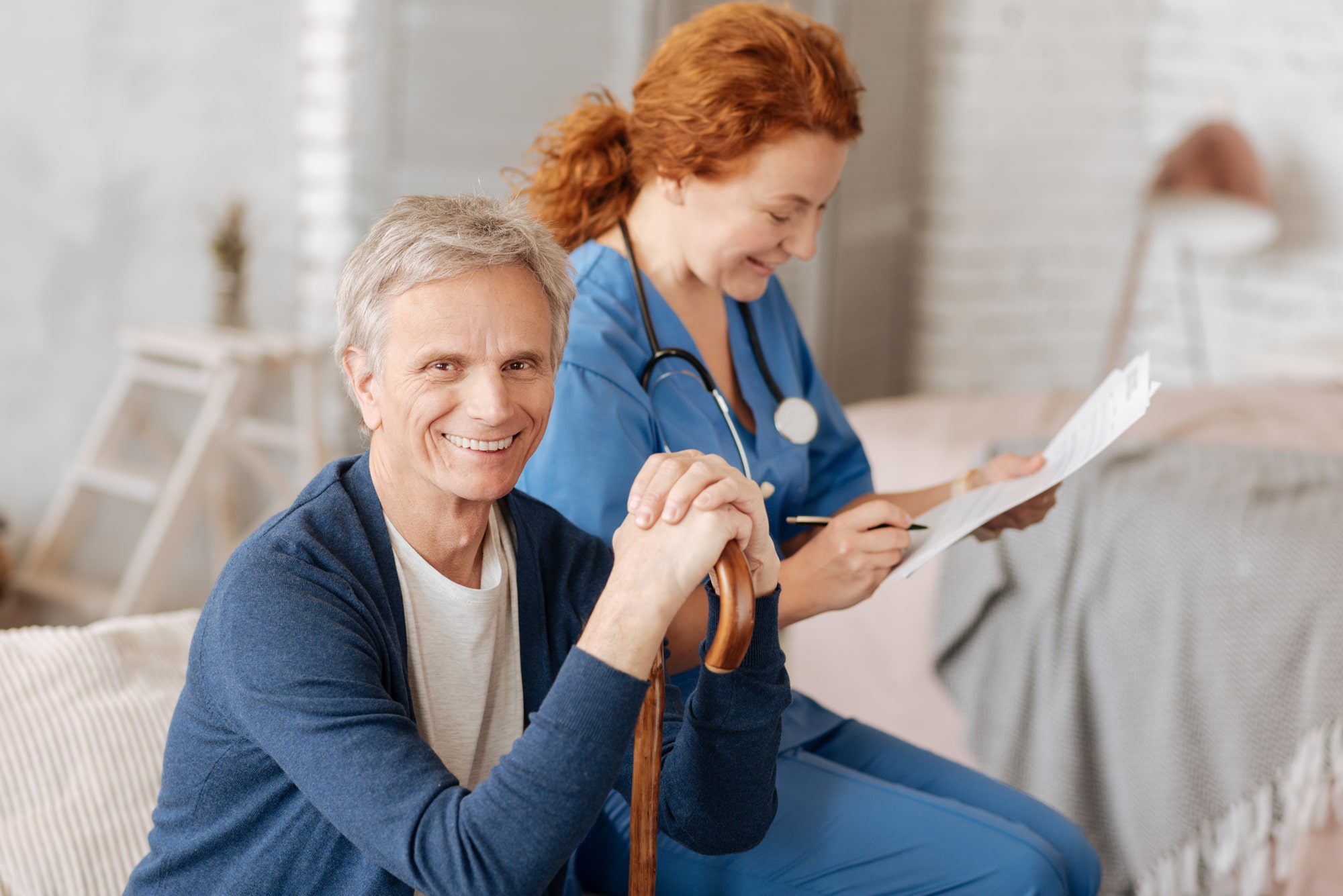 Image of a smiling senior man with a cane sitting while a medical professional takes a needs assessment.